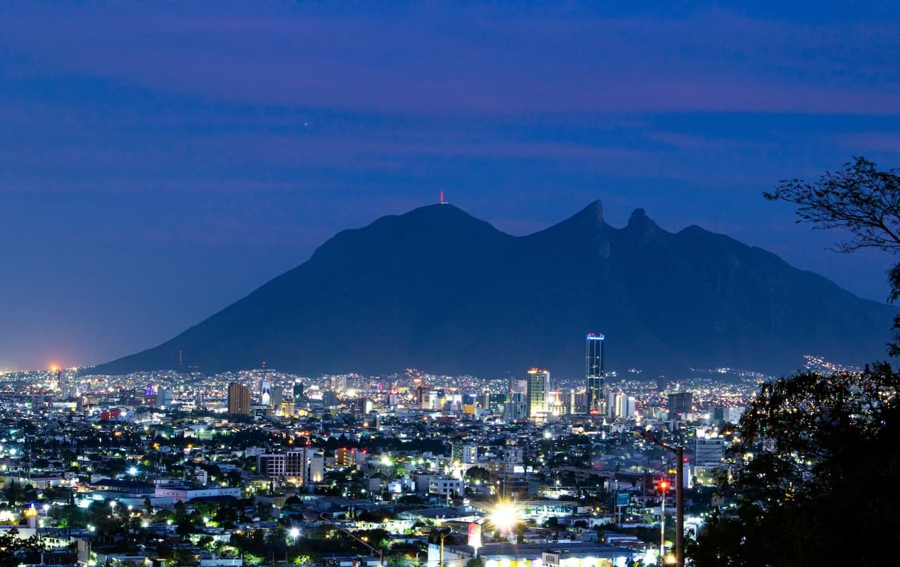 Vista nocturna de una ciudad con montañas al fondo, iluminada por luces urbanas y en un cielo despejado.