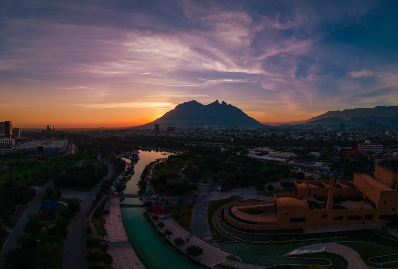 Vista panorámica de la ciudad de Monterrey, México, con la Sierra Madre al fondo y un río iluminado por el atardecer.