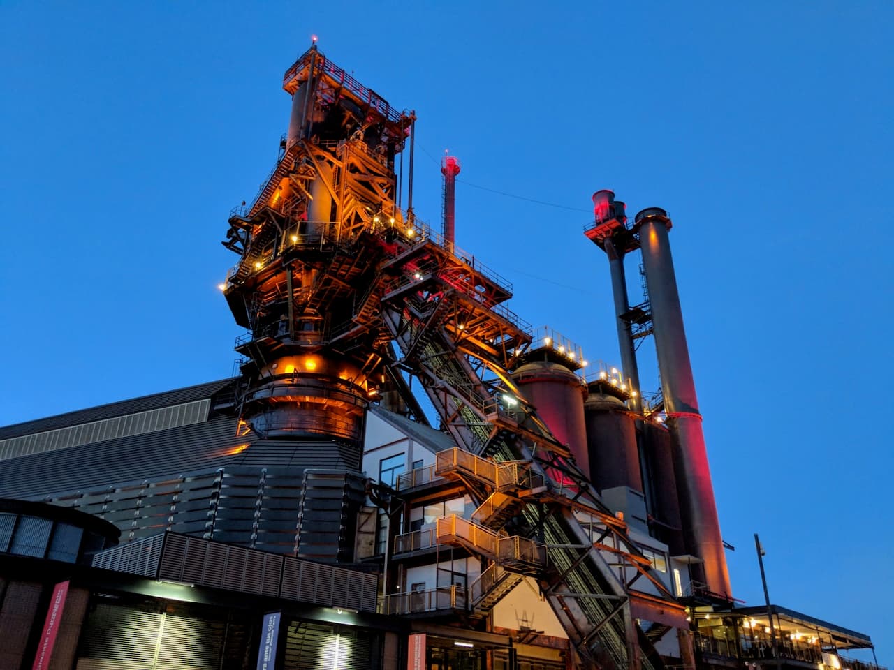 Estructura industrial iluminada durante la noche, con chimeneas y escaleras visibles, bajo un cielo azul profundo.