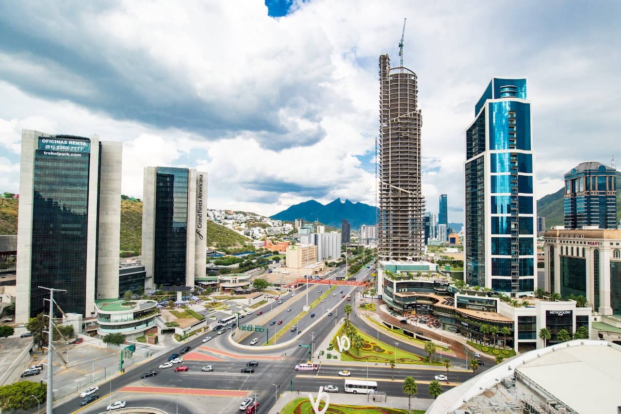 Vista panorámica de una moderna área urbana con edificios altos, una construcción en proceso y montañas al fondo, en un día nublado.