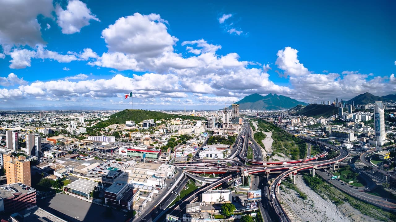 Vista panorámica de una ciudad con un gran infraestructura urbana, montañas al fondo y un cielo azul con nubes. Se observa una gran bandera de México y diversas avenidas que cruzan la zona.