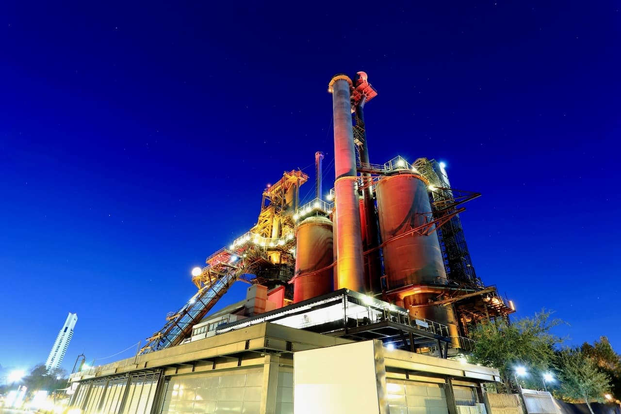 Planta industrial iluminada en un cielo nocturno, con estructuras metálicas y silos en primer plano.